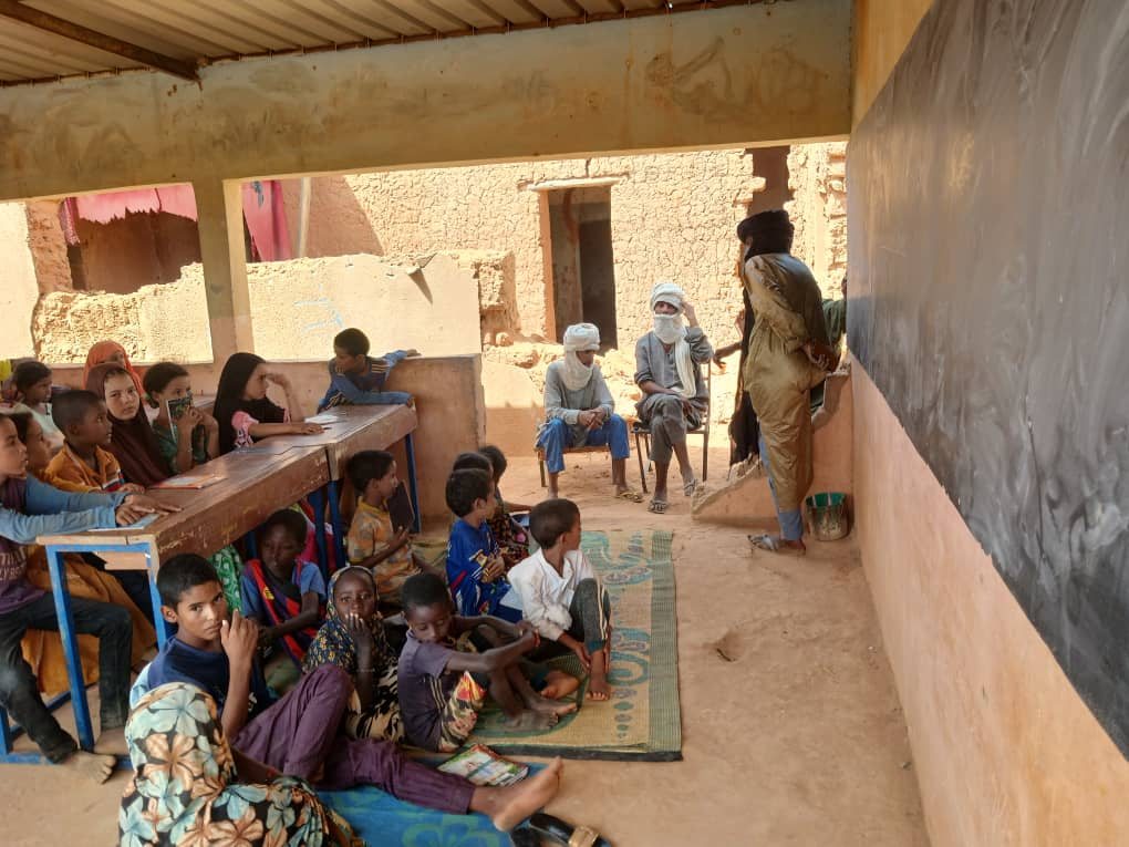 A class under a sheet-metal shelter, facing the blackboard.